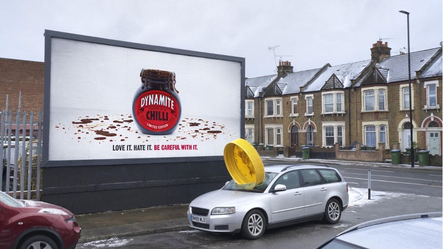 An advert for Marmite with a giant jar lid on top of a car parked in front of the sign